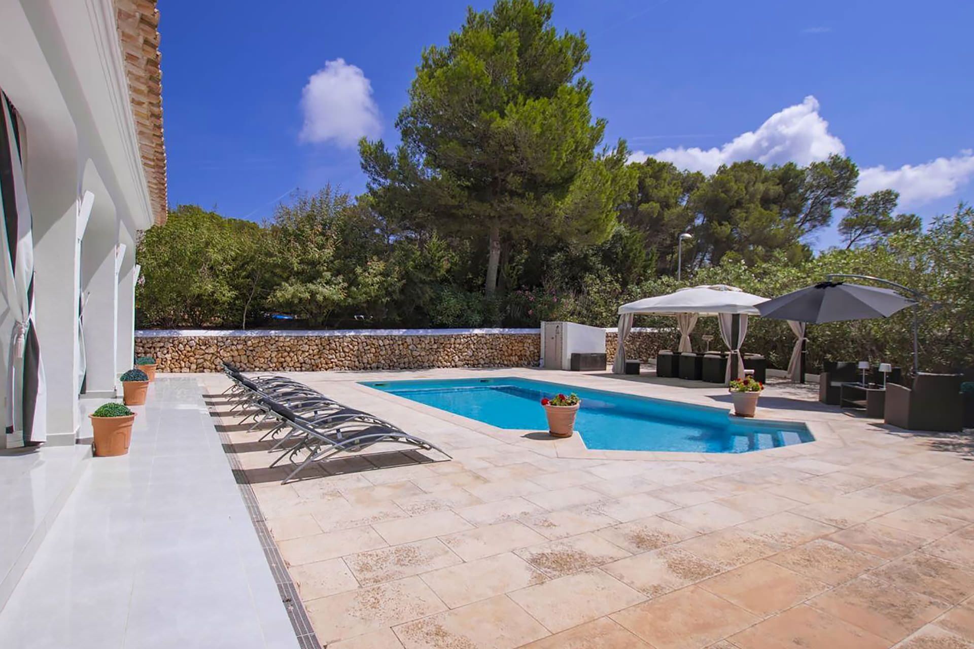 A swimming pool with lounge chairs and a covered seating area, surrounded by trees and a stone wall.