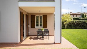 A covered patio with a table and chairs, a window with shutters, and a view of a green lawn and buildings in the background. - Apartment MoKi in Moniga del Garda (Moniga del Garda)