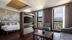 Une chambre d’hôtel comprenant un lit, un téléviseur fixé sur un panneau en bois, un canapé, une table basse et de grandes fenêtres donnant sur la ville.