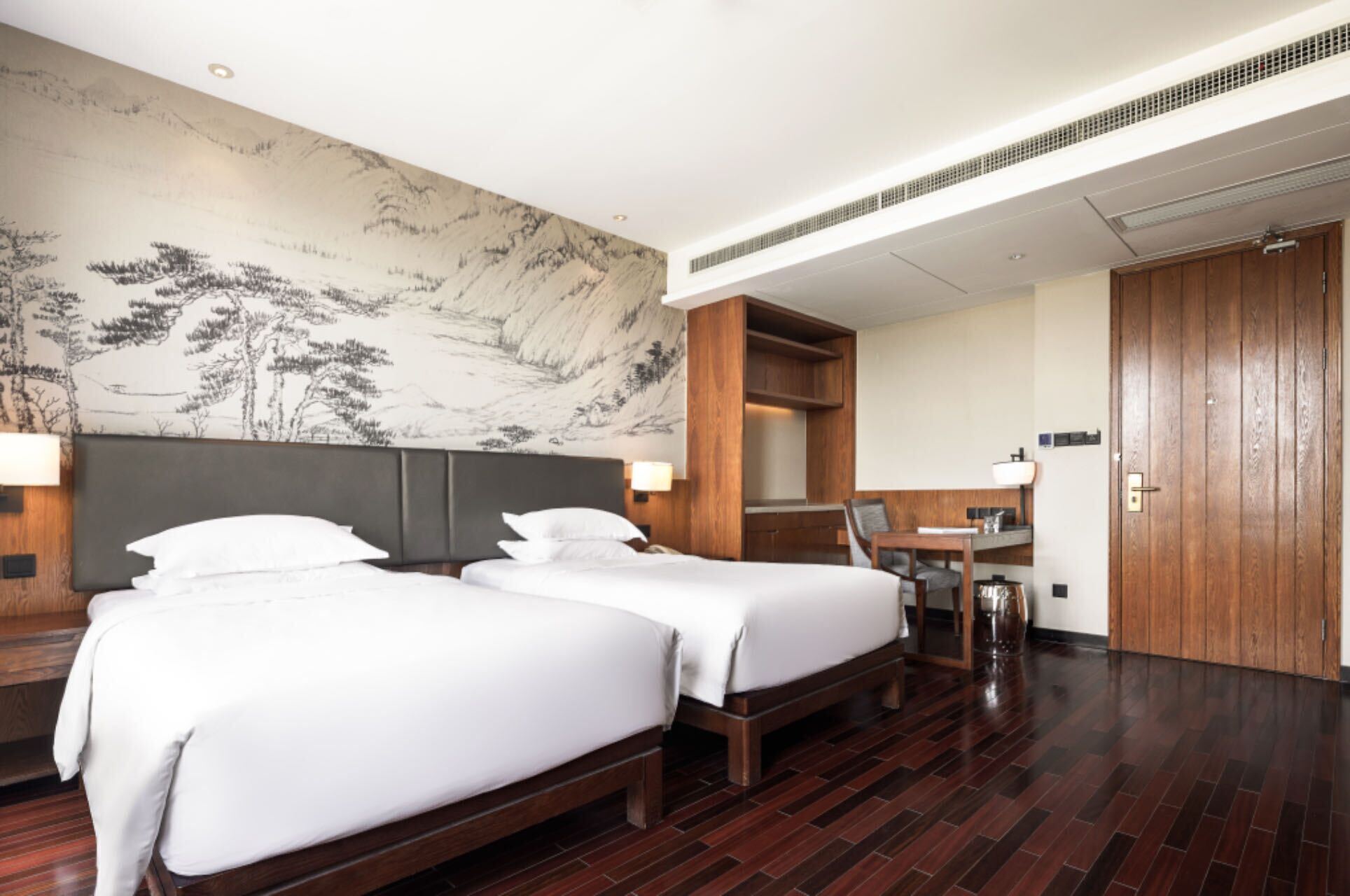 Ein Hotelzimmer mit zwei Betten, einem Schreibtisch, einem Stuhl, einem Holzschrank und einer Wand mit einem traditionellen Gemälde.