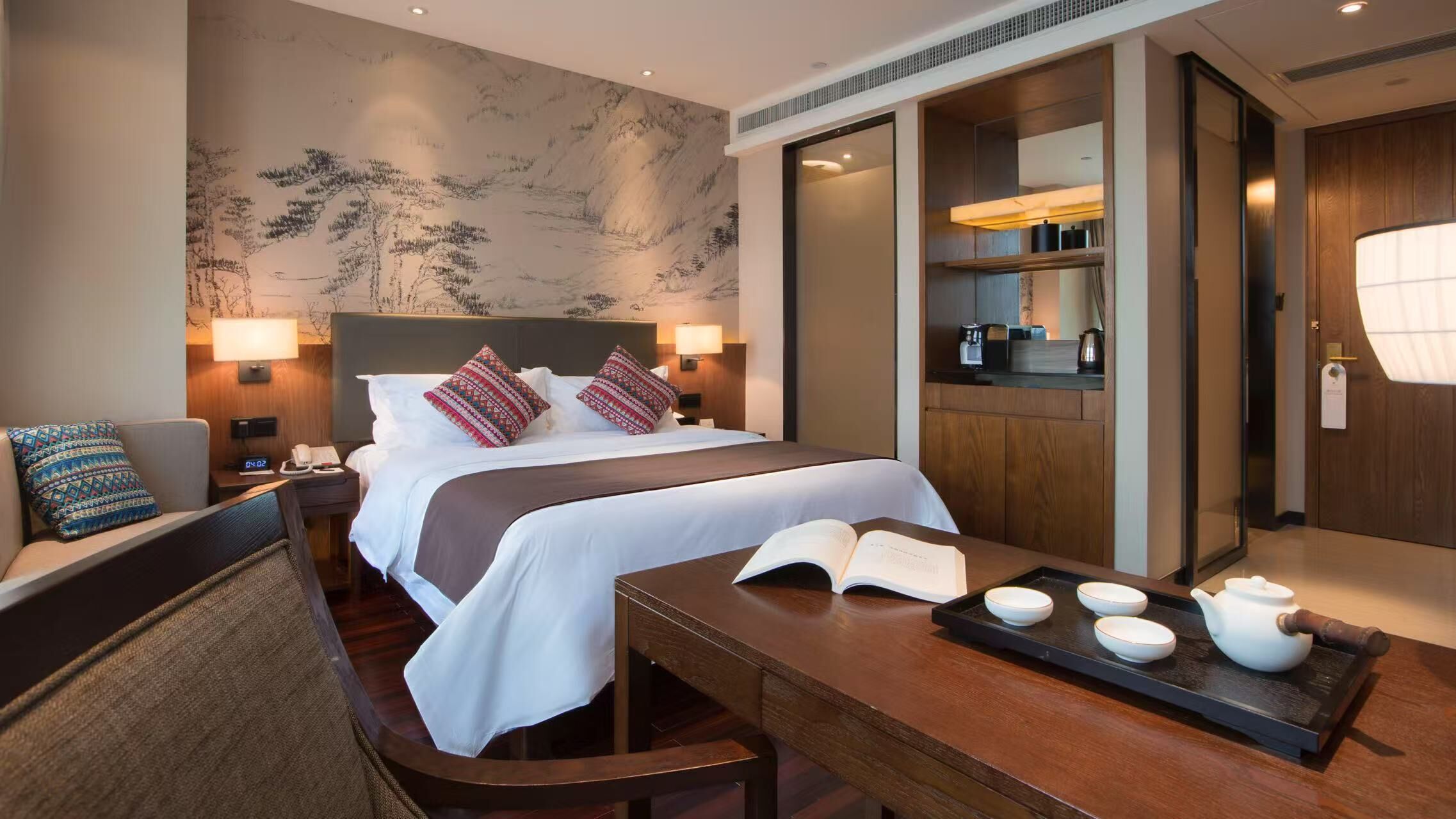 A hotel room with a large bed, bedside lamps, a desk with a tray of teaware, and a view of the hotel's kitchen area.