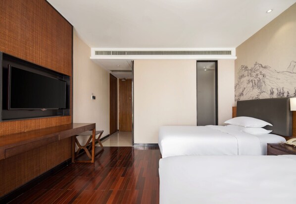 Quarto de hotel com televisão de tela plana na parede, uma escrivaninha de madeira, cama com roupa de cama branca e um quadro na parede.