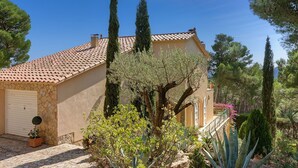 Una casa de estilo mediterráneo con tejado de baldosas, paredes de piedra y un pequeño jardín con diversas plantas y un árbol en maceta.