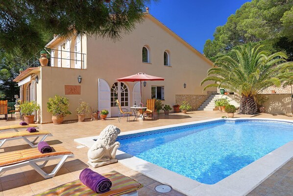 A pool area with lounge chairs and a statue, surrounded by potted plants and a building with a tiled roof.