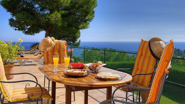 Una mesa de madera con vista al mar, rodeada de sillas a rayas, con una cesta de frutas y una tarta sobre la mesa.