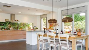 A modern kitchen with a central island, white cabinetry, and a dining area with a wooden table and chairs. - My Rental Homes | Villa Luminosa with private pool and garden (Palafrugell)