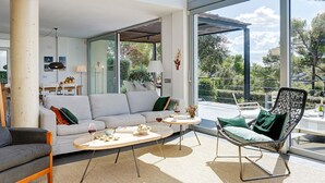 A spacious living area with a large window, a sofa, a coffee table, and a wicker chair. - My Rental Homes | Villa Luminosa with private pool and garden (Palafrugell)