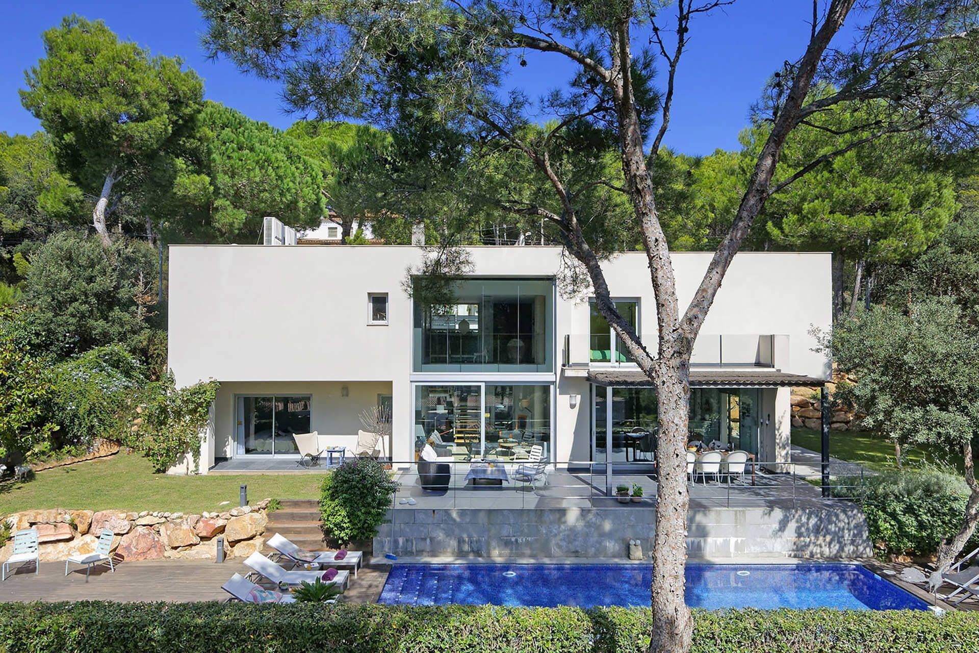 A modern two-story house with a large glass front, a swimming pool, and outdoor seating.