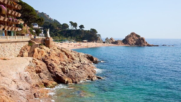 A coastal hotel with a stone facade, a staircase leading to the beach, and a view of the sea and rocky outcrops. - My Rental Homes | Villa Luminosa with private pool and garden (Palafrugell)