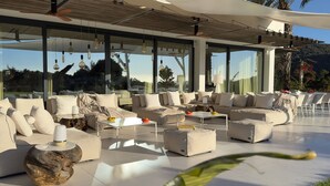A modern outdoor living area with sofas, tables, and a large glass wall offering views of the surrounding landscape.