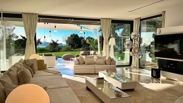 A modern living room with a large sofa, a glass coffee table, and a TV. The room has large windows offering a view of a garden and mountains.