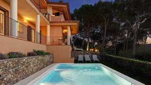 Una piscina con sillones de descanso y un muro de piedra, rodeada de árboles y un edificio con balcones.