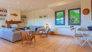 Een ruime woonkamer met een bank, stoelen, een open haard, een boekenkast en een groot raam met uitzicht op groen.