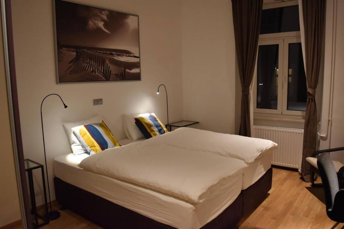 Ein Schlafzimmer mit einem großen Bett, zwei Nachttischen, einem Beistelltisch, einem Fenster mit Vorhängen und einem Wandbild.