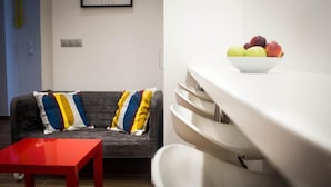 Un salon moderne avec un canapé gris, des coussins colorés, une table rouge et un coin cuisine blanc agrémenté d’un panier de fruits.