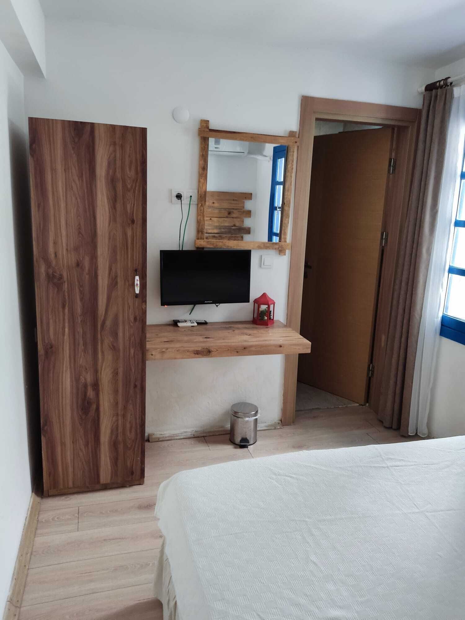 A bedroom with a wooden wardrobe, a TV on a wooden stand, a small trash bin, and a bed with a white cover.