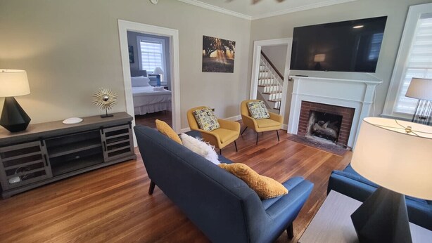 A living room with a blue sofa, yellow armchairs, a fireplace, and a TV mounted above it. - Historic Mayor`s Manor | 2 King Bds • Fenced Yard (Sanford)