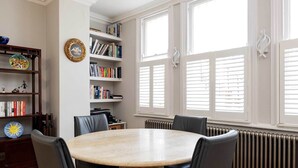 A dining area with a round table, black chairs, a bookshelf, and a radiator. - The Maida Vale Garden Apartment, by Veeve (London)