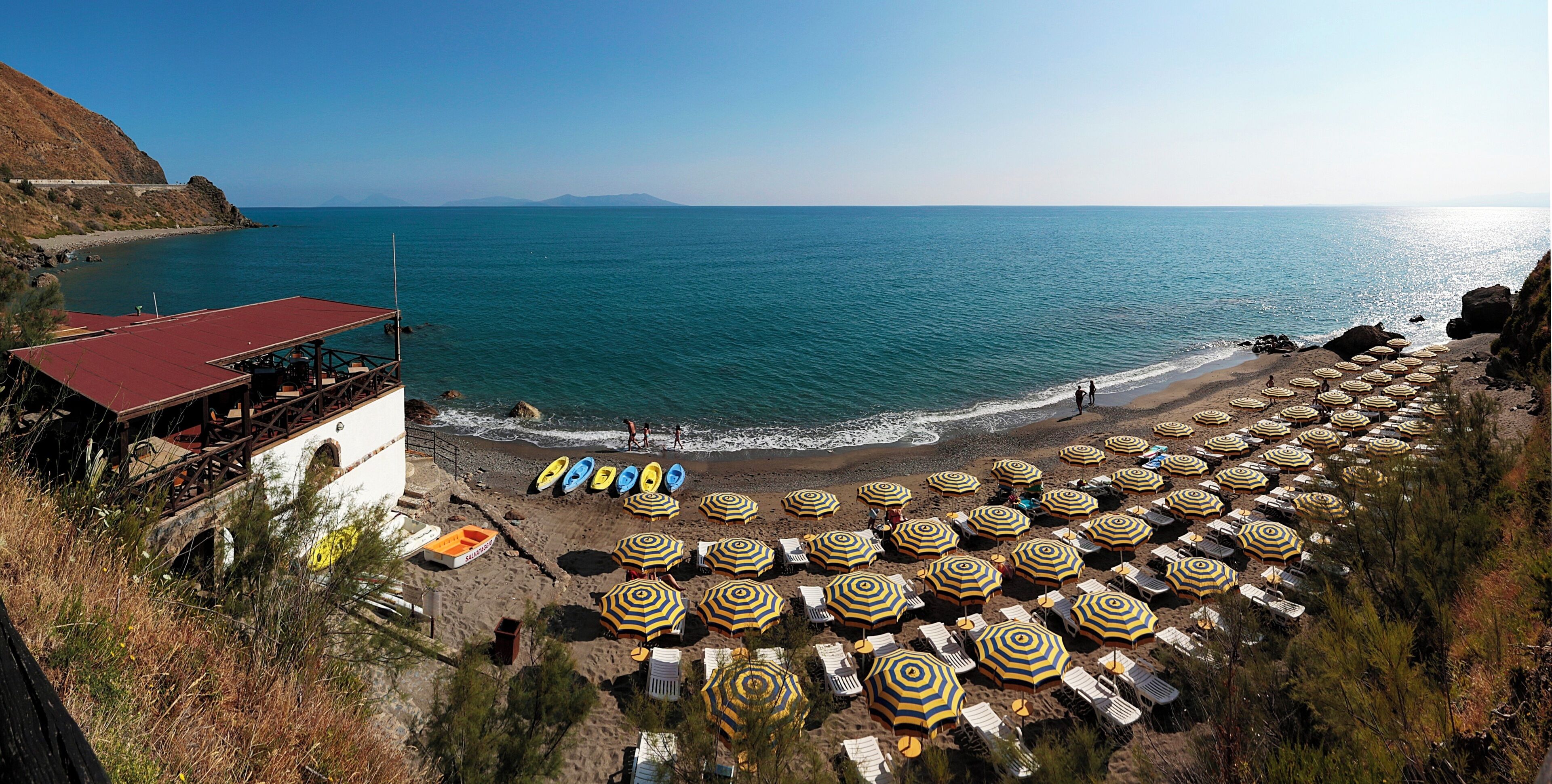 Une plage bordée de rangées de parasols et de chaises longues, un rivage rocheux et une vue sur la mer.