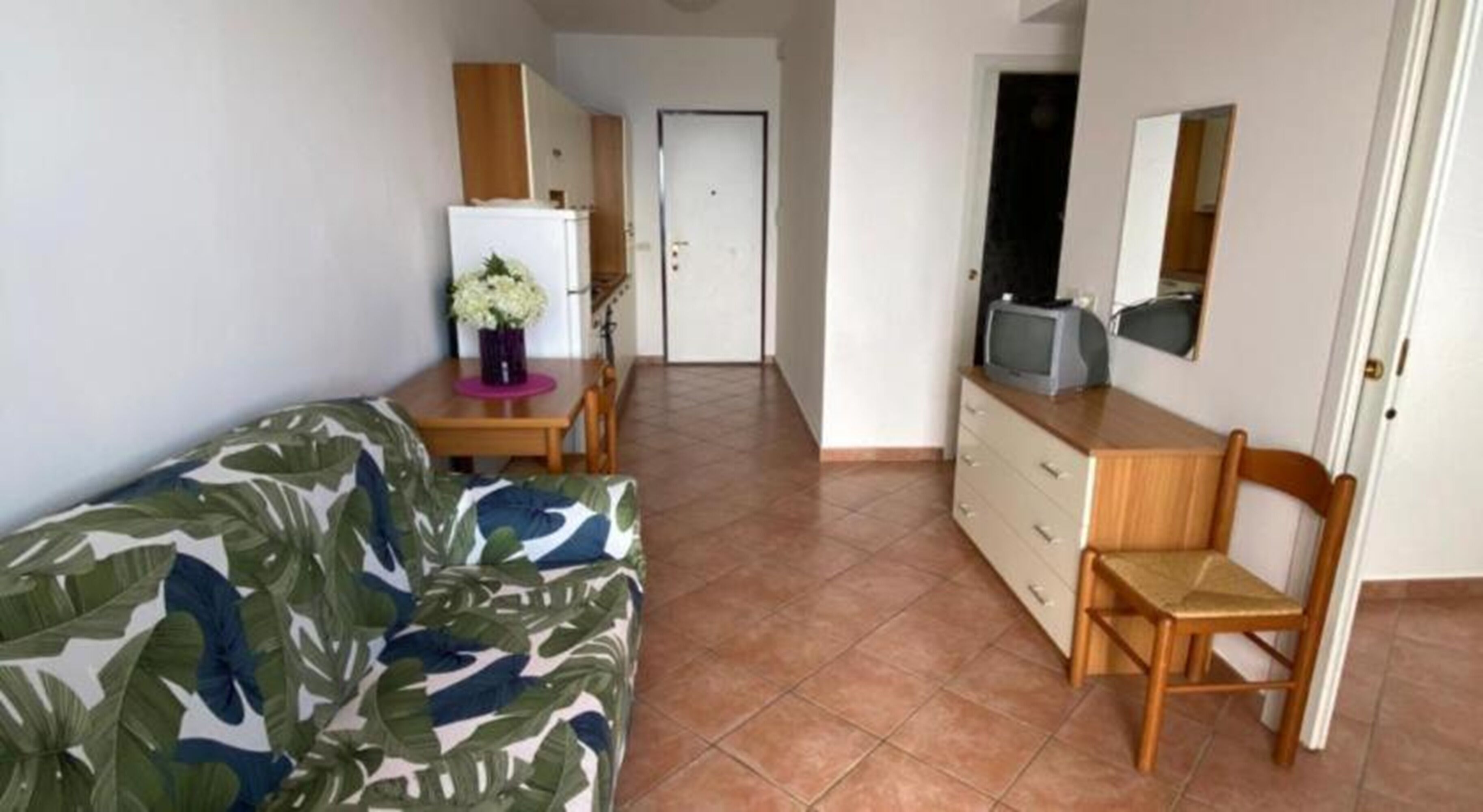 Ein kompakter Wohnraum mit einer Couch, einem kleinen Esstisch mit Stühlen, einem Kühlschrank, einer Holzkommode und einem Fernseher.