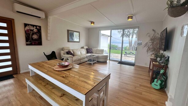 A spacious living room with a wooden dining table, a beige sofa, and a television mounted on the wall.