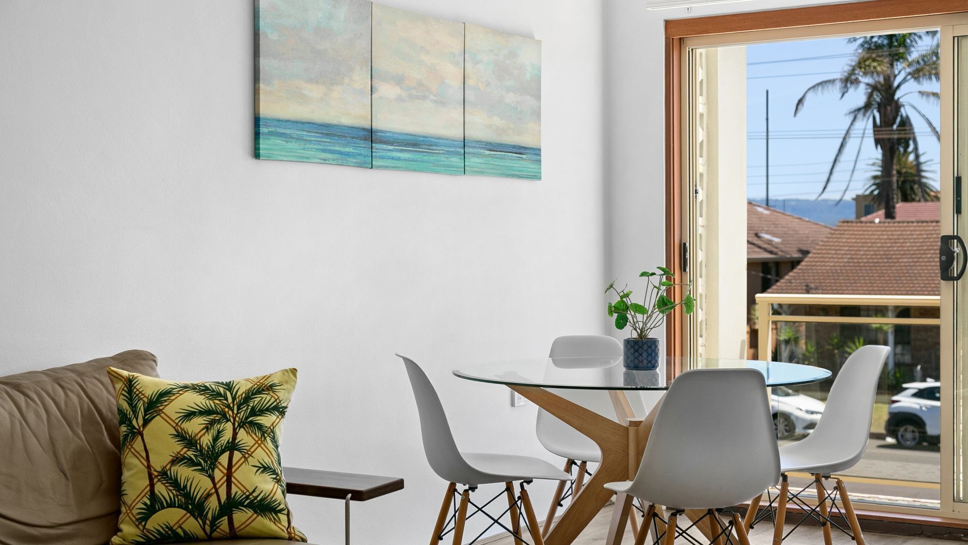A modern room with a glass table, white chairs, a sofa with a palm tree pillow, and a painting of a beach scene.