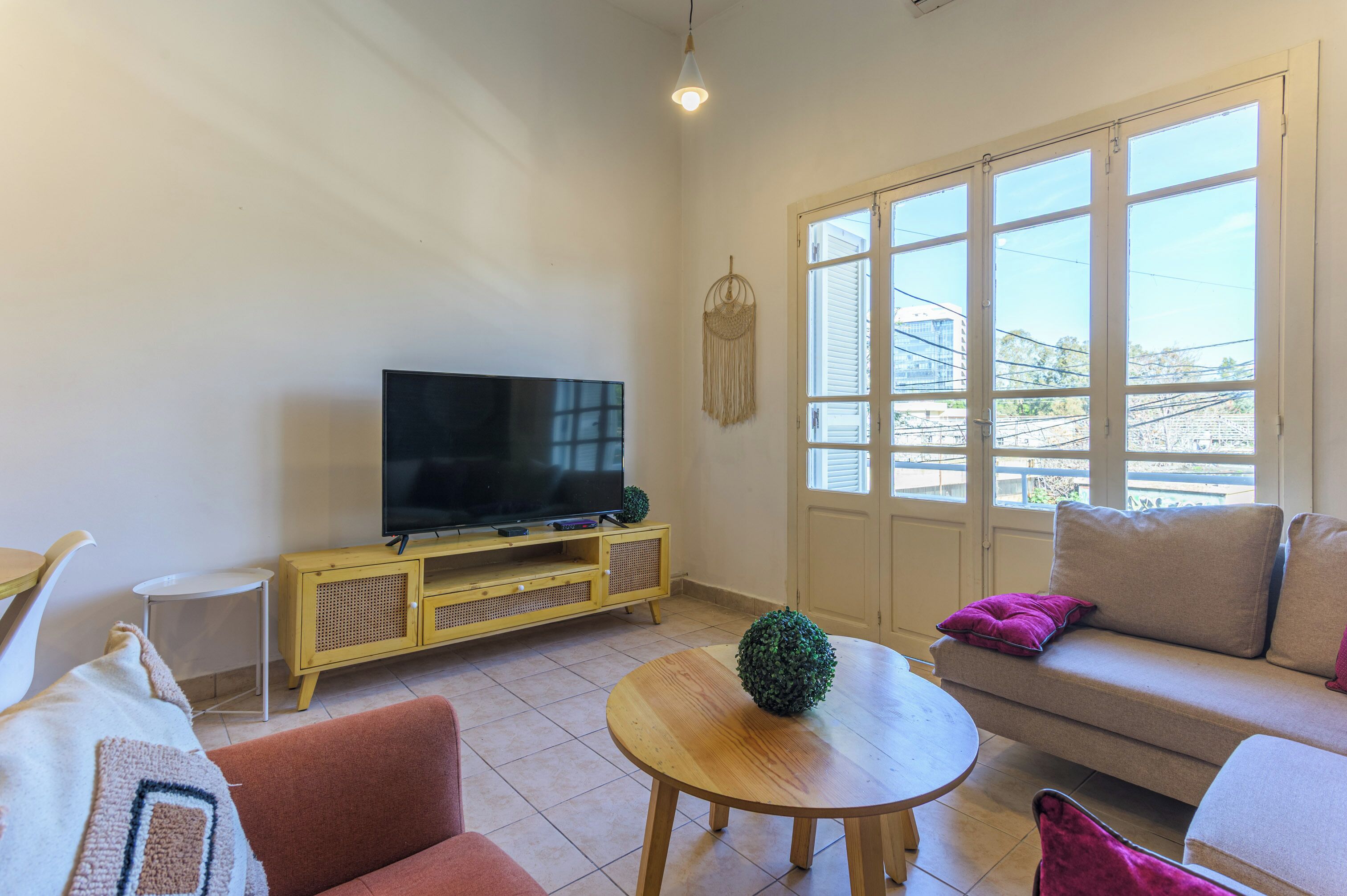 Una sala de estar con televisor sobre un mueble de madera, una mesa redonda de madera, un sofá y un amplio ventanal con vistas a edificios y líneas eléctricas.