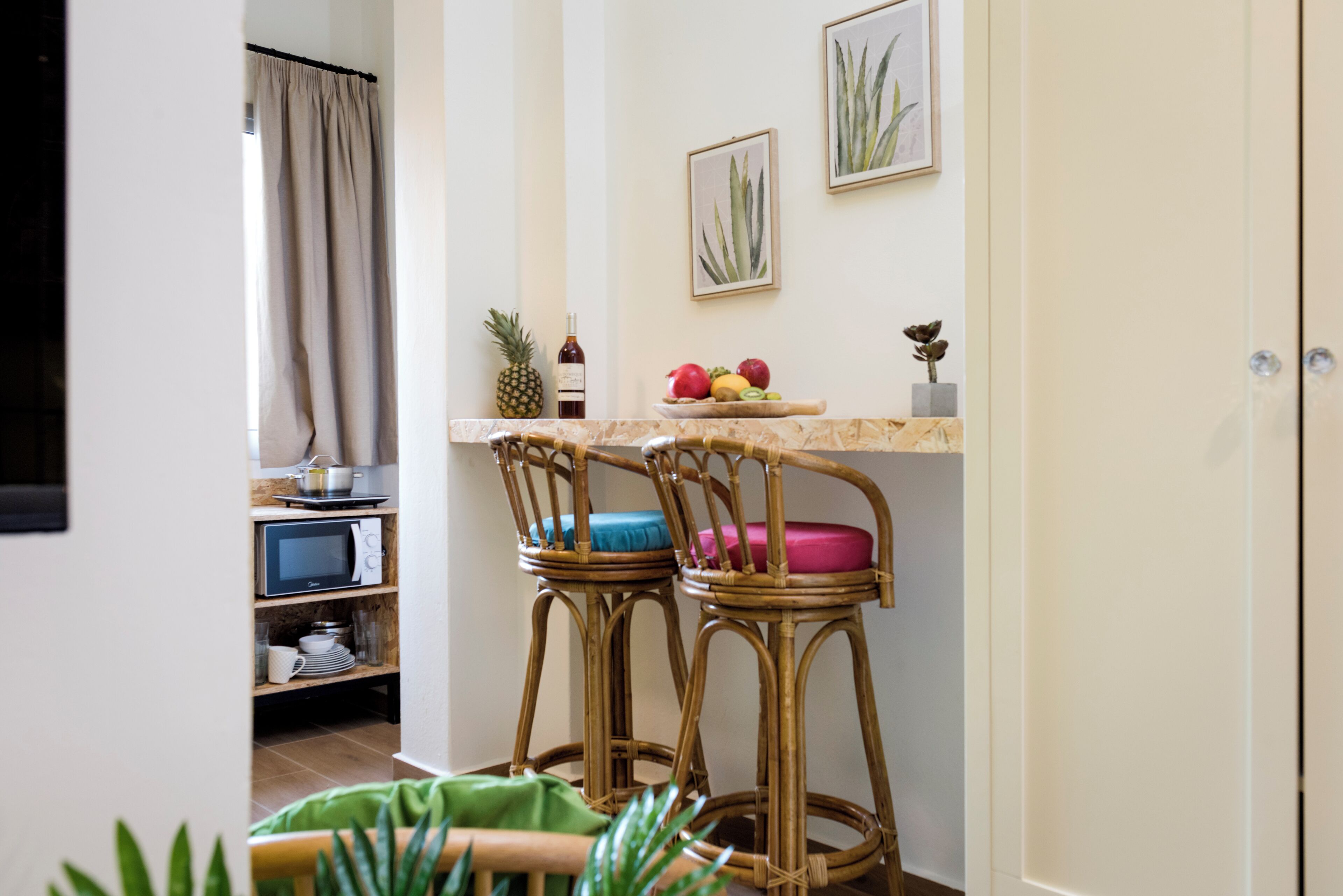 Una zona de cocina con dos taburetes de mimbre, un microondas, una estantería con utensilios de cocina y un frutero en la encimera.