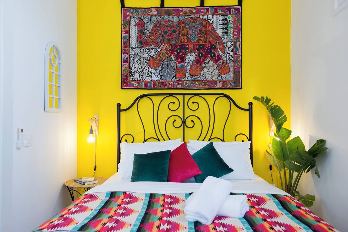 Ein Bett mit einer farbenfrohen Steude, eine Wand mit gelb-weißer Musterung und ein großes Elefantenwandteppich.