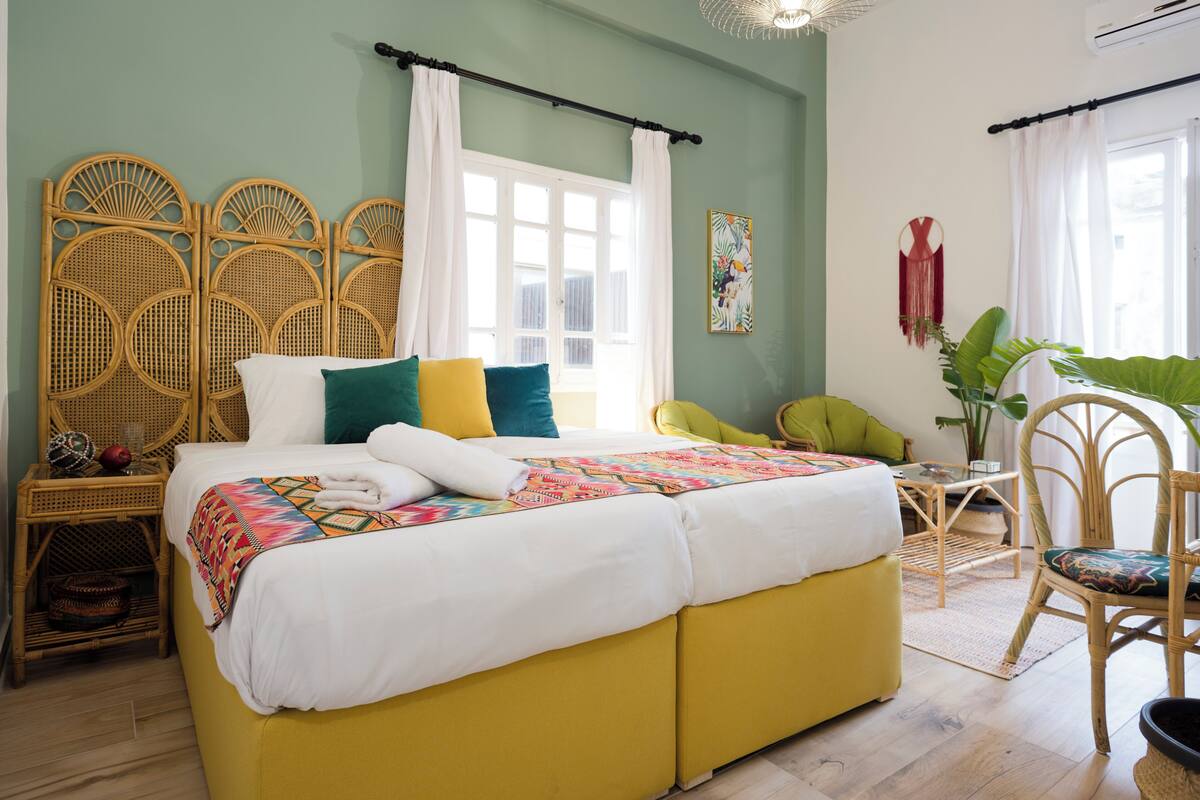 Ein Bett mit einer farbenfrohen Steude, ein gelber Hocker, Korbmöbel und ein großes Fenster mit weißen Vorhängen.