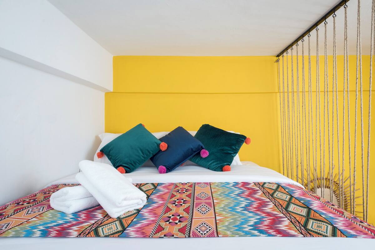 Ein Bett mit einer farbenfrohen, gemusterten Tagesdecke, weißen Kissen und einer gelben Wand.