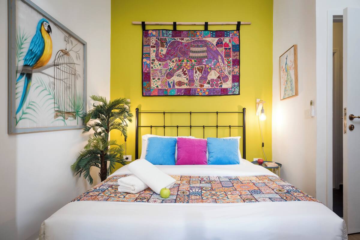 Ein Bett mit farbenfroher Bettwäsche, eine Wand mit Wandteppich mit Vogel und Elefant, und eine Zimmerpflanze neben dem Bett.