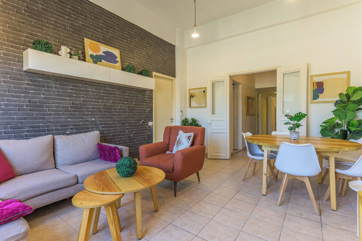 Ein modernes Wohnzimmer mit Backsteinwand, einer Couch, einem Couchtisch, einem roten Sessel und einem Essbereich mit Holztisch und Stühlen.