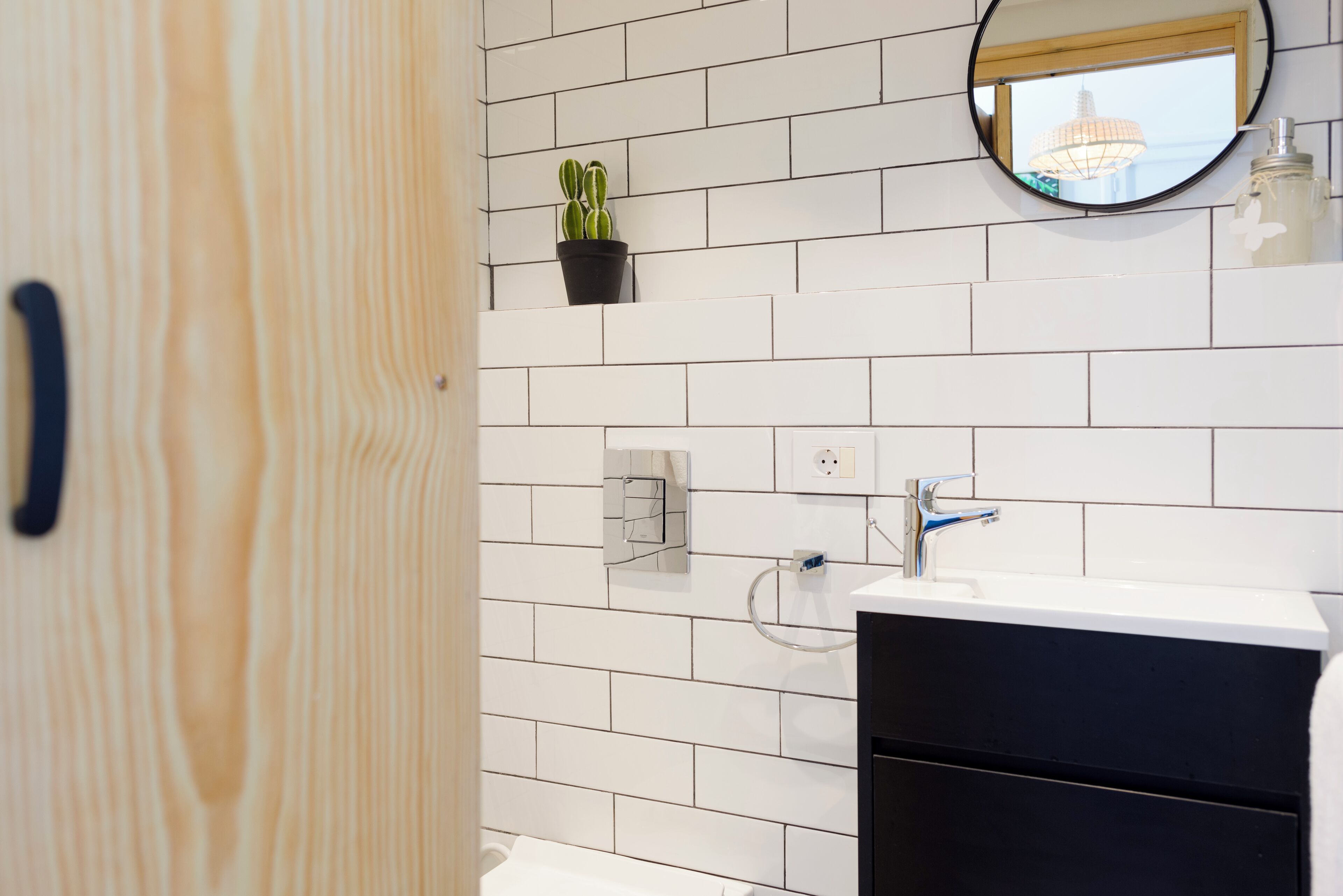 A bathroom with white tiled walls, a black cabinet, a white sink, a mirror, and a potted plant.
