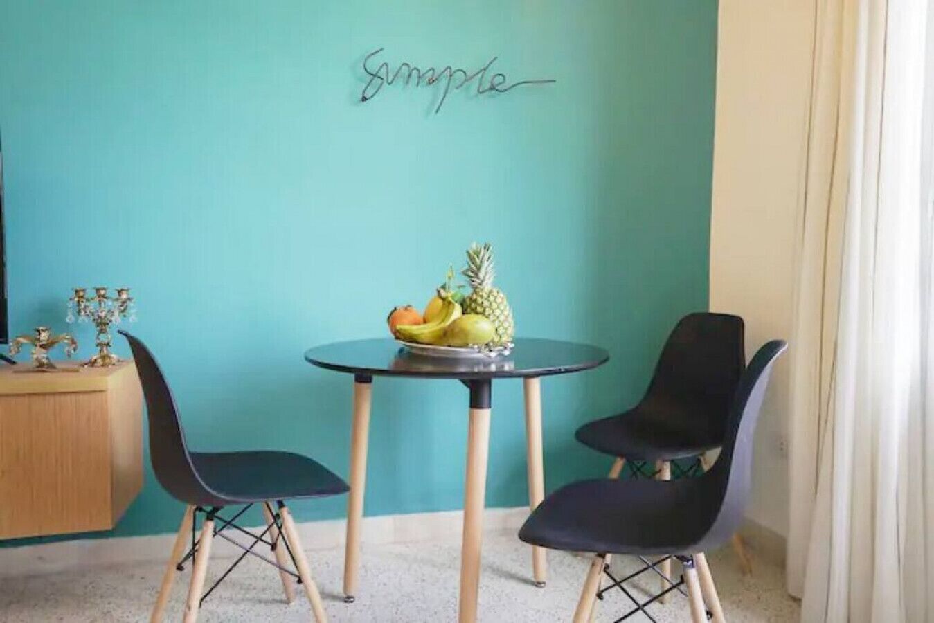A teal-colored wall with a "Summy" sign, a round table with a fruit bowl, two black chairs, and a wooden cabinet.