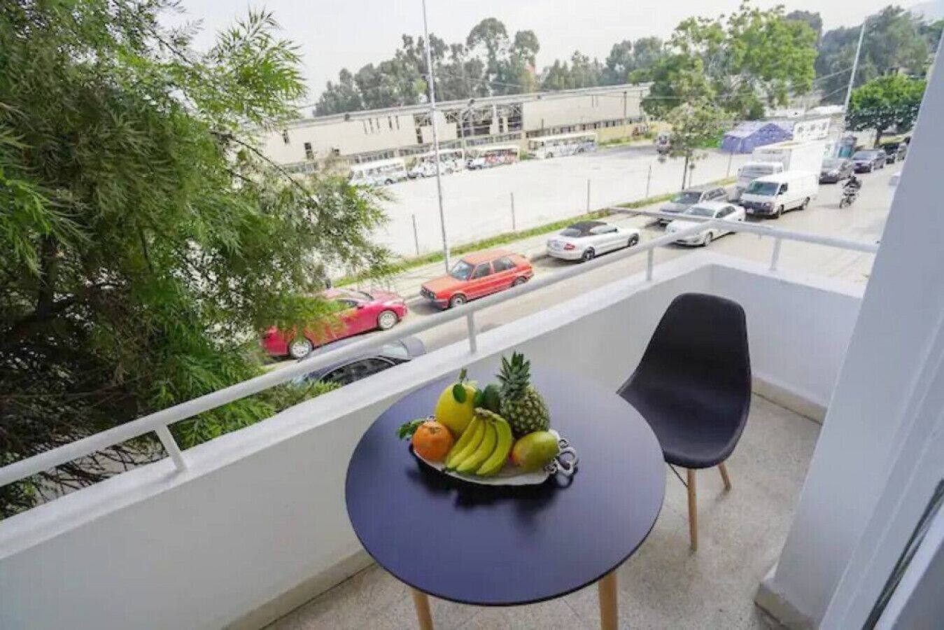 Un balcón con una mesa y sillas, un frutero y vista a un estacionamiento con coches y un edificio al fondo.