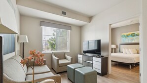 A modern hotel room with a sofa, armchair, ottoman, TV, and a bed.