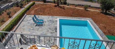 A terrace with a pool, dining table with food and drinks, and a view of the sea and hills.