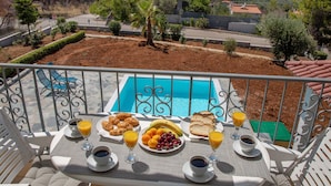 A balcony with a table set for breakfast overlooking a swimming pool, with a view of a modern building and a hilly landscape. - Villa with S. Pool Theologos by GHH (Lokroi)