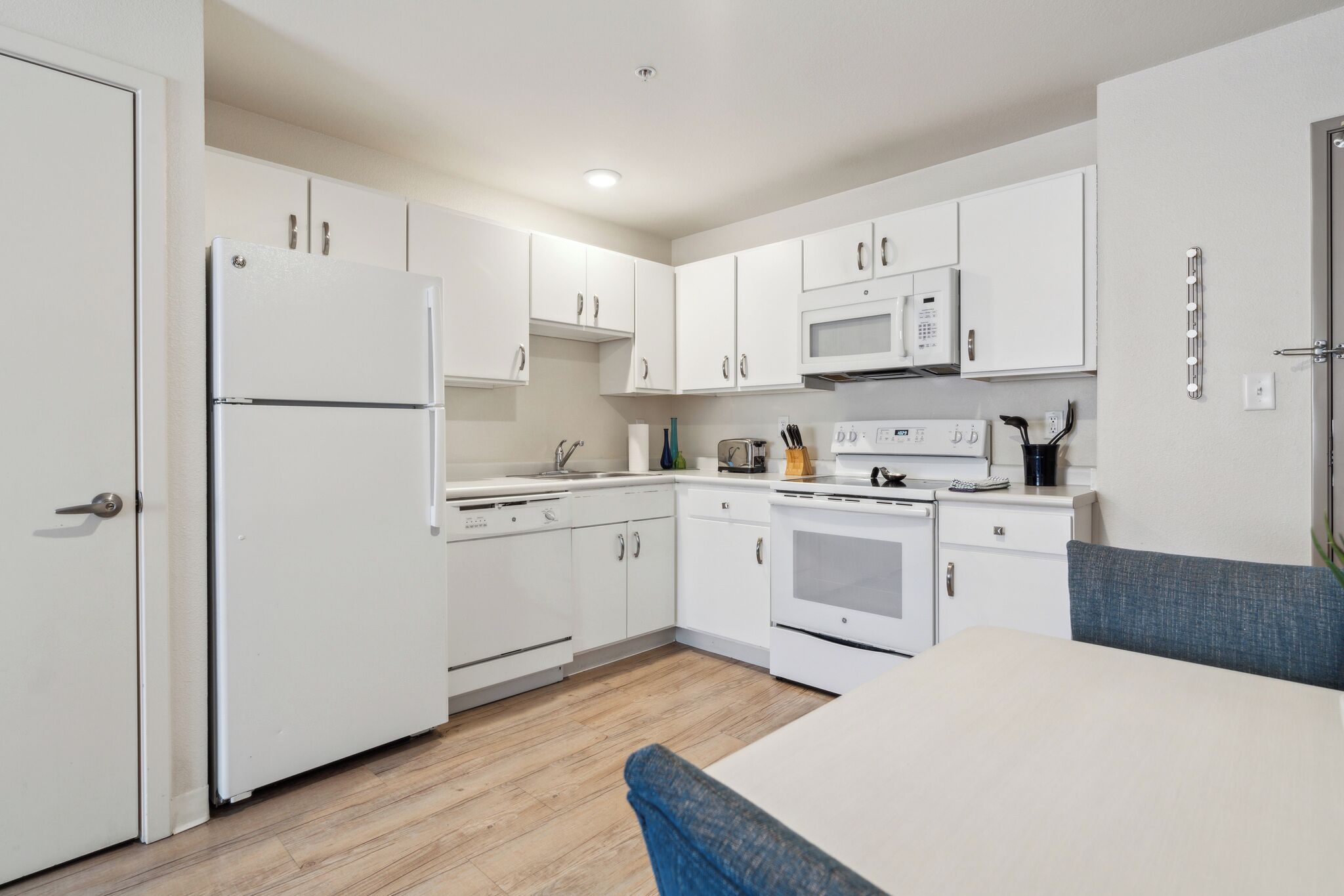A compact kitchen with white cabinets, a microwave, a refrigerator, a toaster, and a dining area with a table and a blue chair.