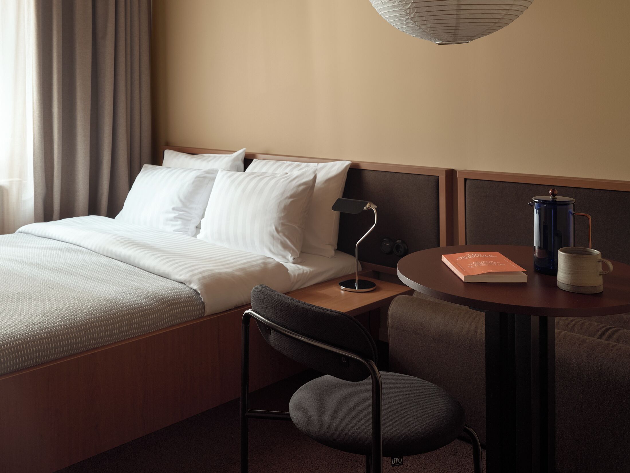 A hotel room with a bed, a desk with a lamp, a coffee maker, and a chair.