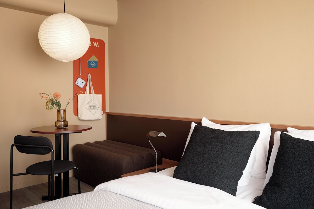 A hotel room with a bed, a desk with a chair, a hanging lamp, and a wall-mounted sign.
