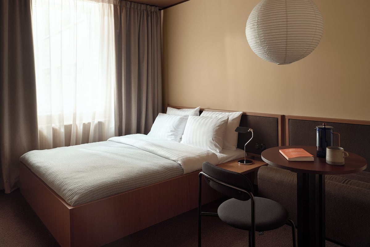 A hotel room with a bed, a desk with a chair, a lamp, a book, a coffee maker, and a hanging light fixture.