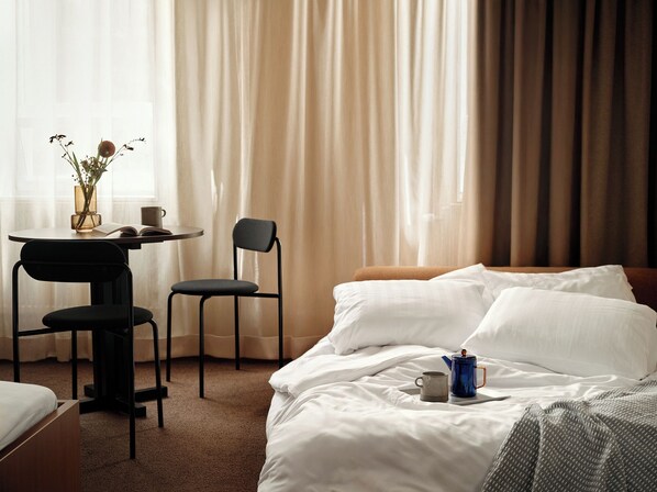 Une chambre d’hôtel avec un lit, une petite table sur laquelle est posé un vase de fleurs, une chaise et une cafetière sur le lit.