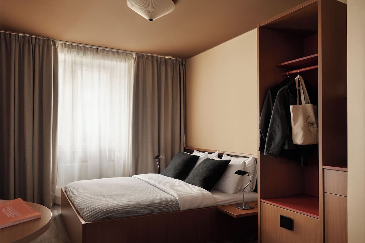 A hotel room with a bed, bedside table, lamp, and a wardrobe with hanging clothes.