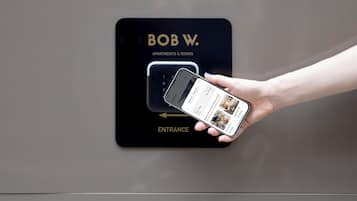 Uma mão segurando um smartphone com a interface de um aplicativo de hotel, apontando para uma placa que diz 'BOB W. APARTMENTS & ROOMS ENTRANCE'.