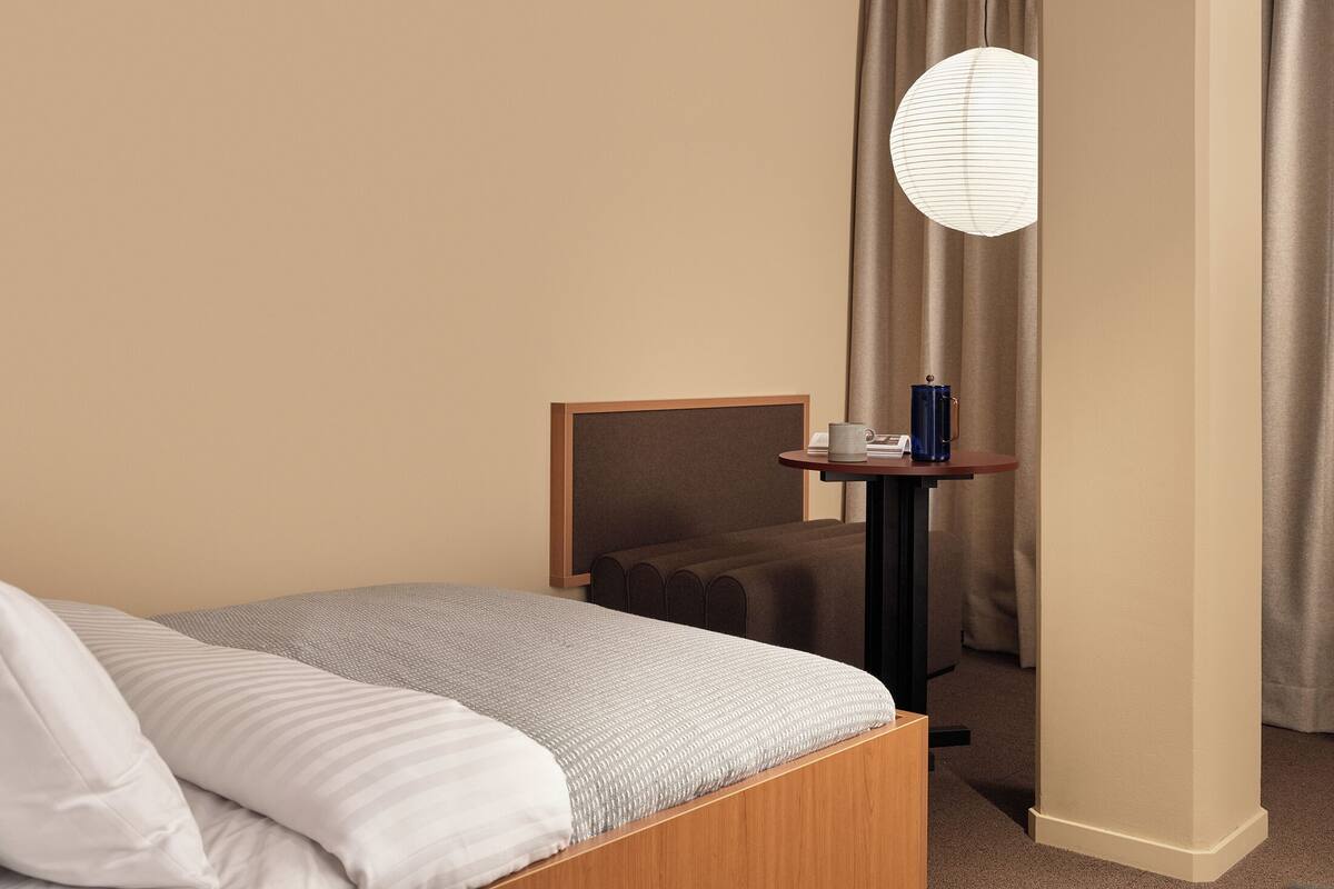 A hotel room with a bed, a bedside table with a lamp, and a view of the outside through a window with curtains.