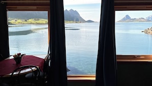 A room with a large window offering a view of a lake and mountains. There is a table with chairs and a vase of flowers. - Fjordcamp AS (Straumsjøen)