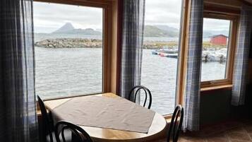 Een eethoek met een tafel en stoelen, grote ramen met gordijnen en uitzicht op water en bergen.