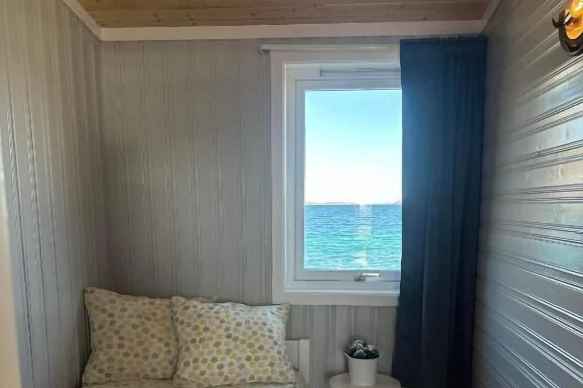 A small, narrow room with a bed, a window with a view of the sea, and a small table with a plant.